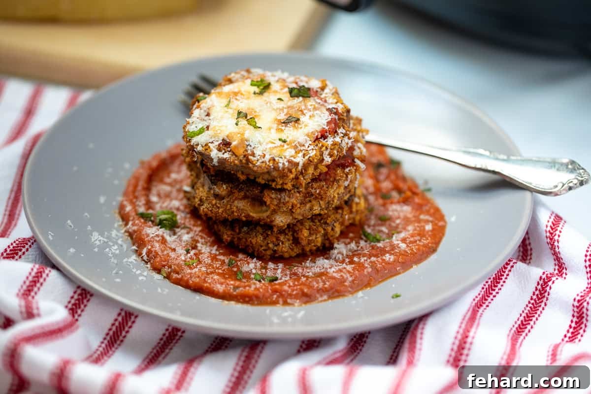 Artfully stacked slices of Air Fryer Eggplant Parmesan on a gray plate, with an air fryer visible in the blurred background, showcasing the dish's irresistible appeal.
