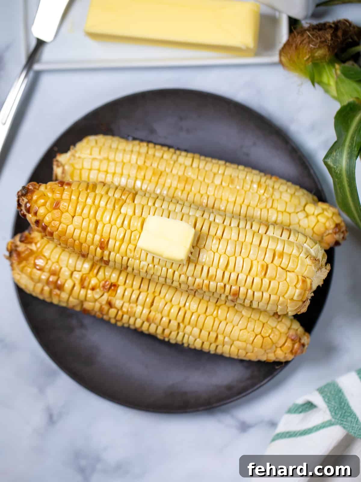 Air Fryer Corn on the Cob 3 ears of air fryer corn on a black plate topped with a pat of butter.