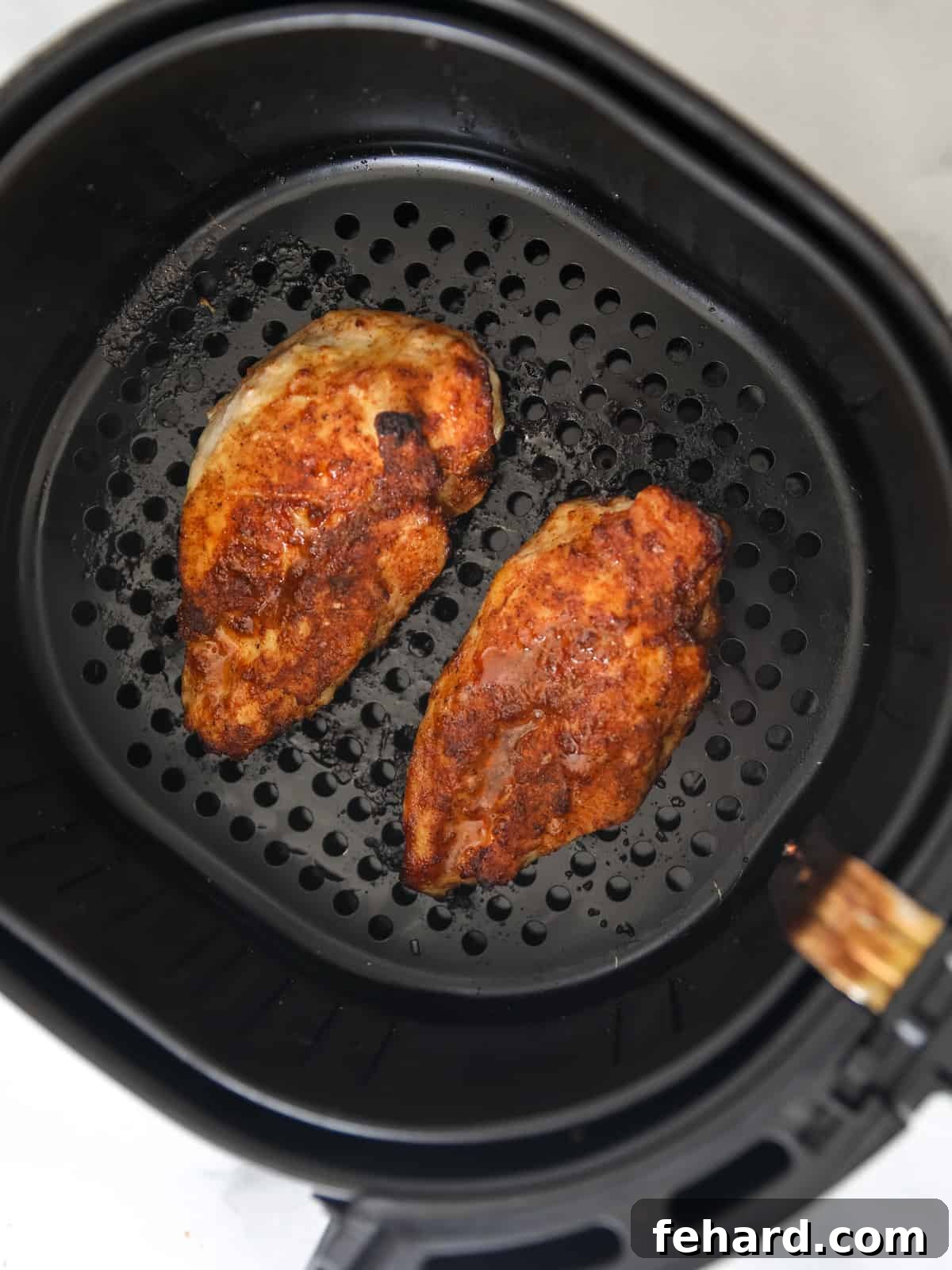Two perfectly golden brown, spice-rubbed chicken breasts cooking in an air fryer basket, showing a crispy texture.