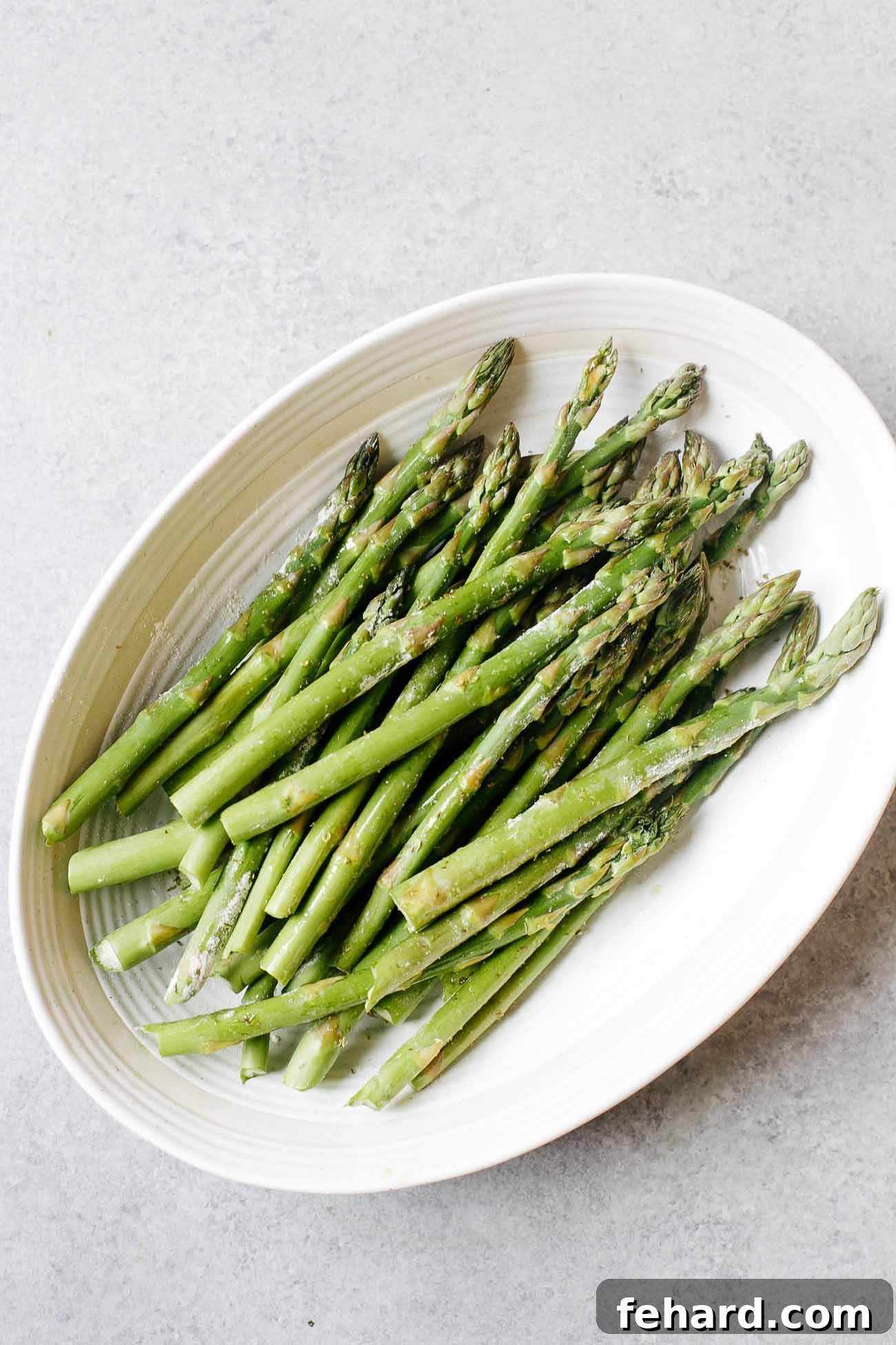 Freshly trimmed asparagus spears neatly arranged in a white casserole dish, ready for seasoning and cooking.