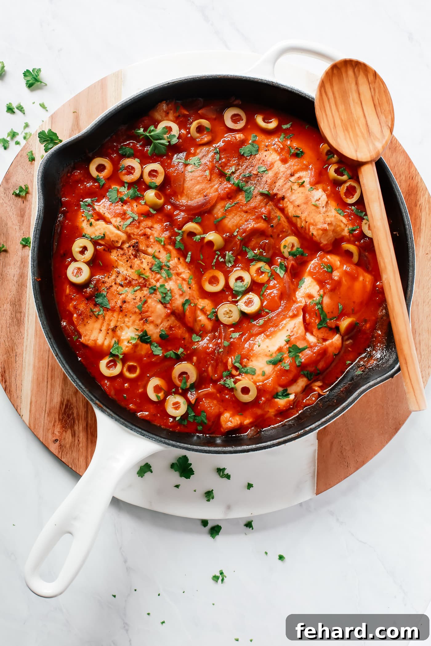 Overhead view of a white skillet containing perfectly cooked tilapia fillets nestled in a vibrant tomato and onion sauce, garnished with fresh herbs.