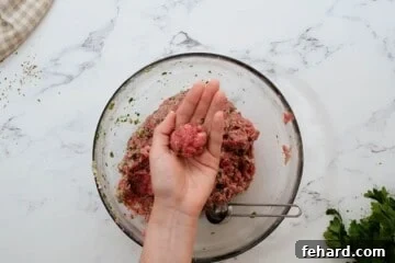 Hand showing rolled meatball with tablespoon measuring spoon in bowl with meatball mixture.