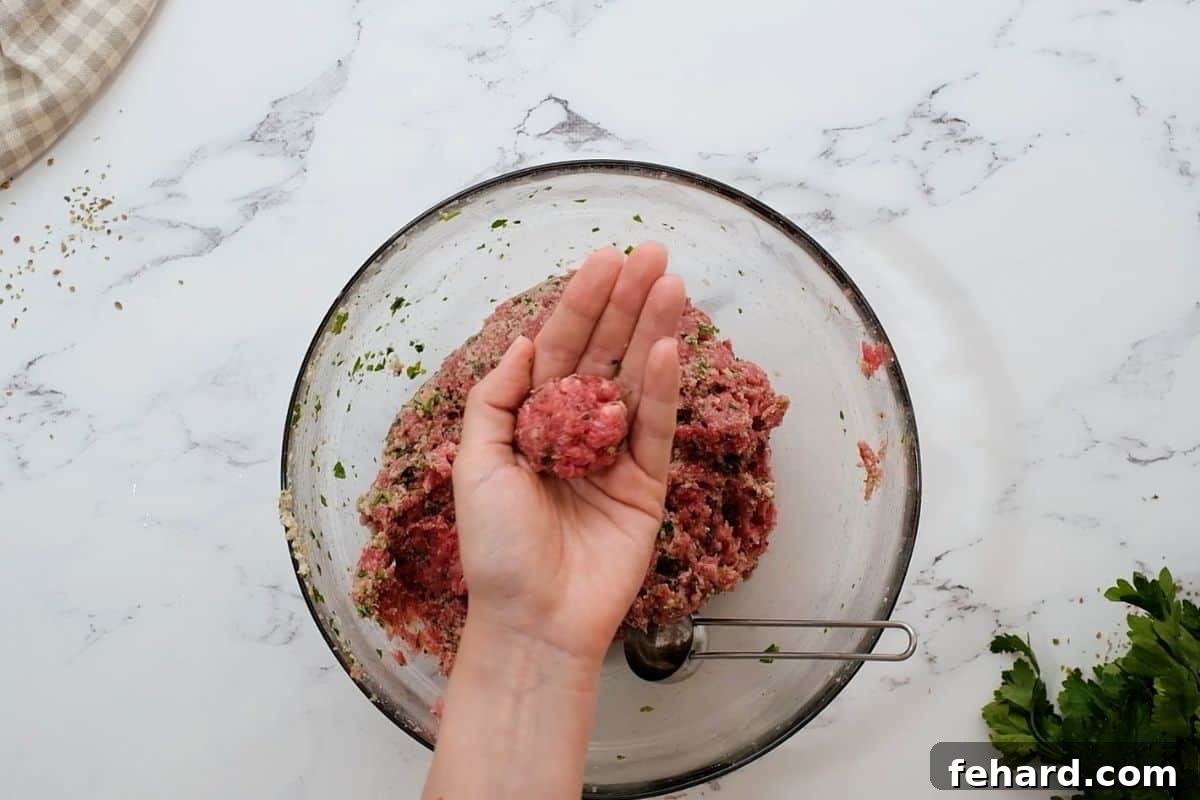 A hand demonstrating how to roll a meatball, with a tablespoon measuring spoon in a bowl of meatball mixture.