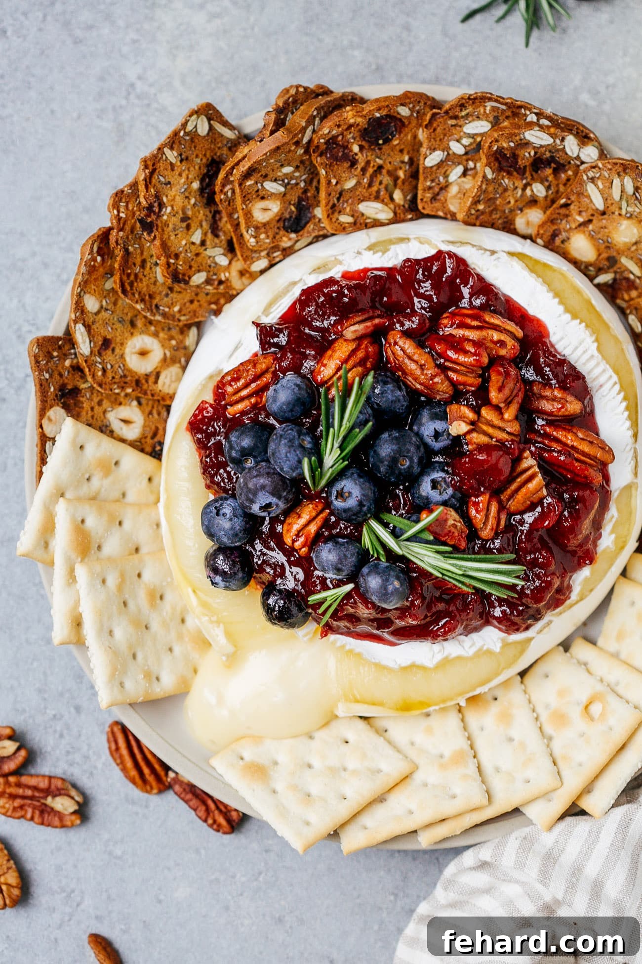 Overhead view of a baked brie with cranberry sauce served with crackers and fresh rosemary.