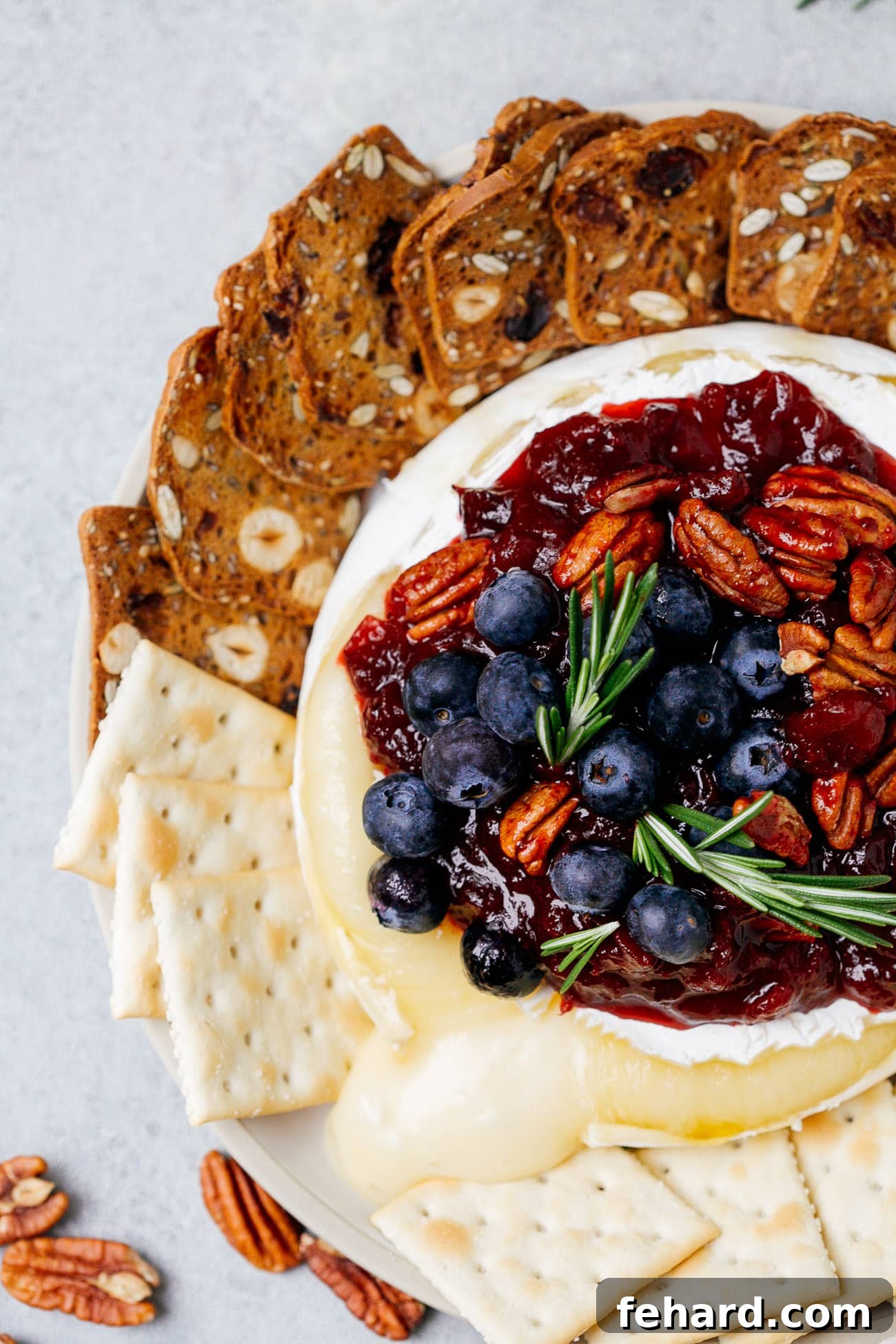 Overhead view of a baked brie with cranberry sauce with pecans, blueberries, and rosemary on top, ready to be served.