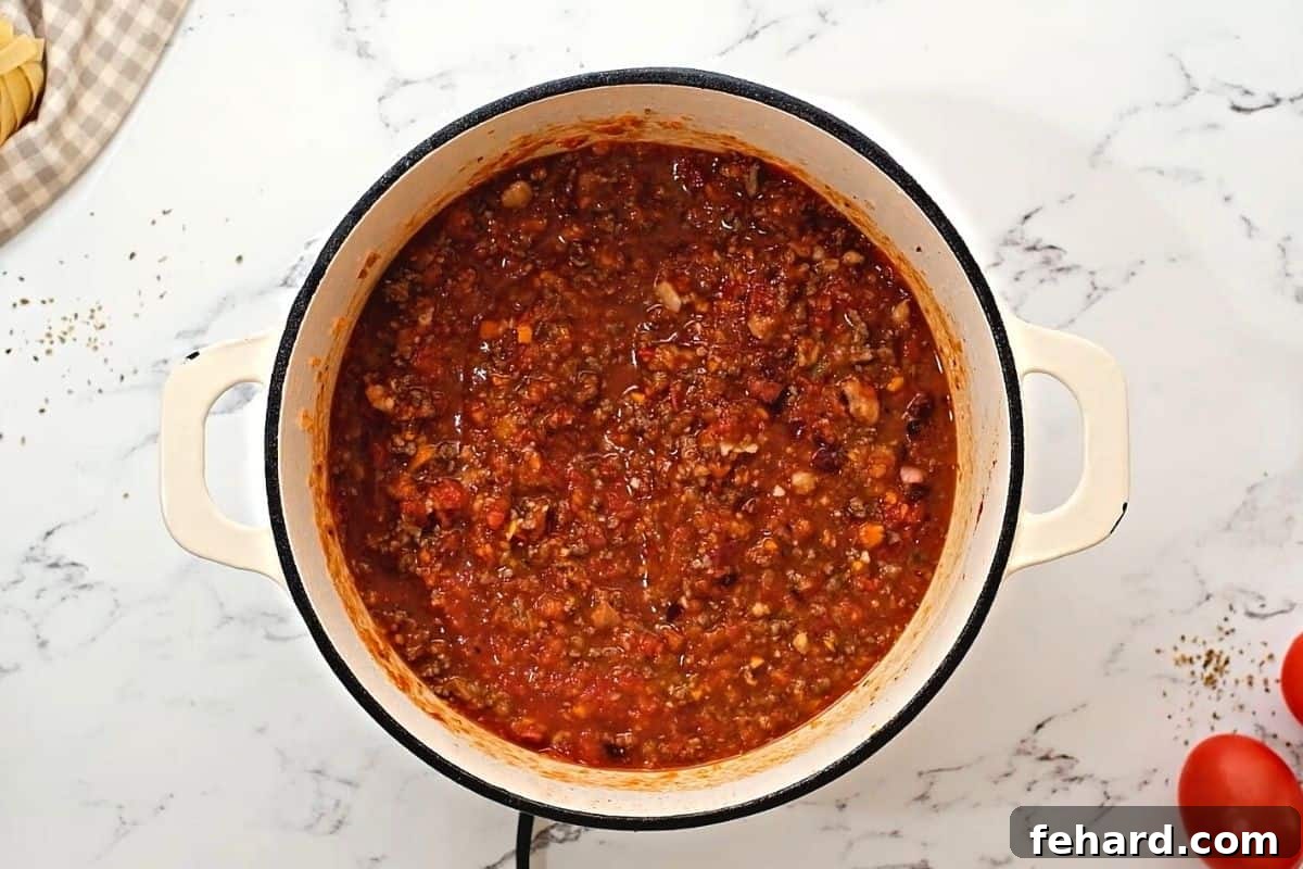 Rich Bolognese sauce gently simmering in a large red Dutch oven, with steam rising, indicating a slow cooking process.
