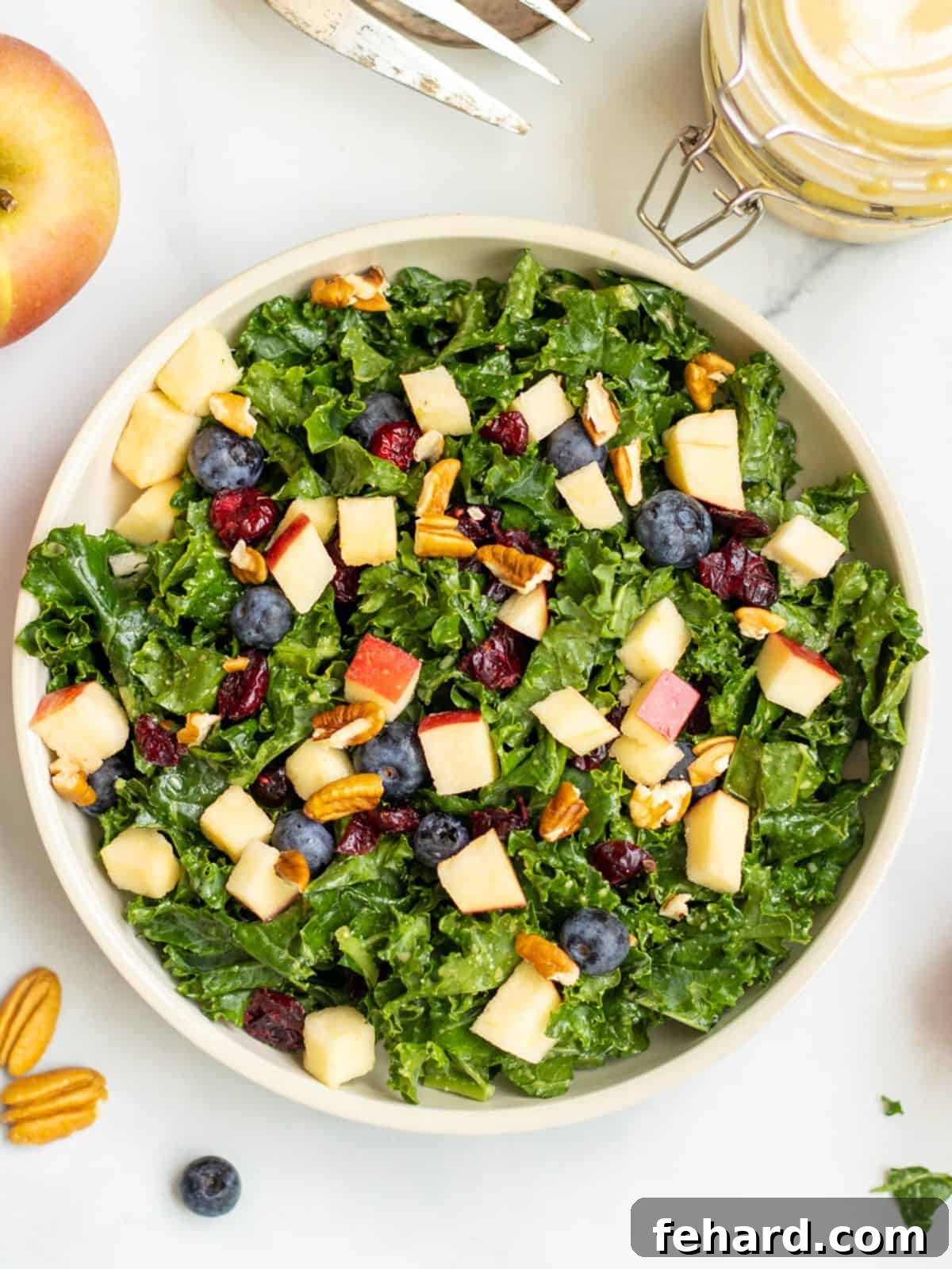 A vibrant Kale Apple Salad in a serving bowl, garnished with pecans and apple slices in the background. The salad features tender kale, bright red cranberries, and golden toasted nuts.