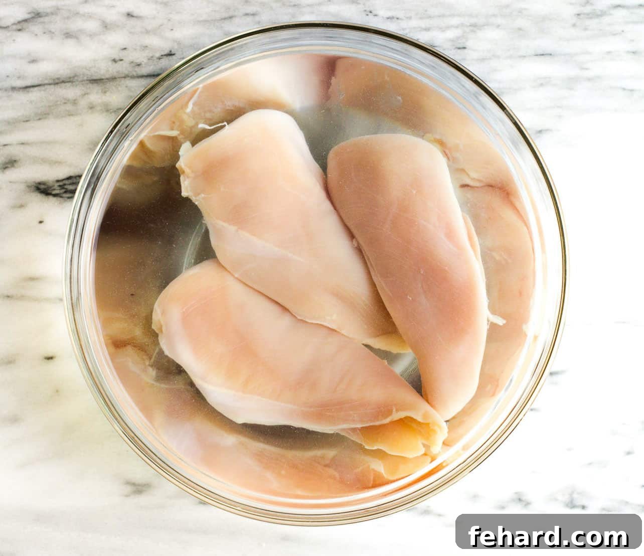 overhead view of raw chicken breast in a bowl with water and salt
