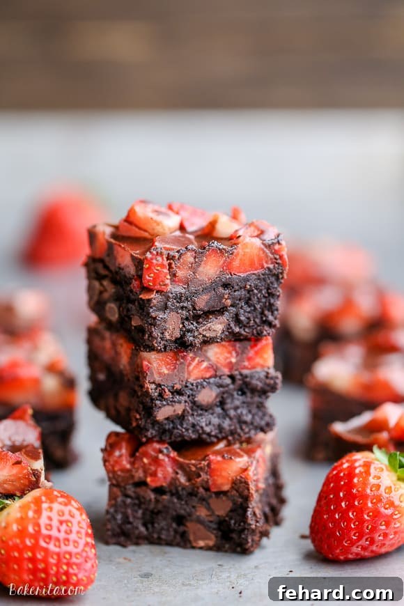 A perfectly stacked trio of chocolate-covered strawberry brownies, with whole, fresh strawberries artfully arranged beside them.