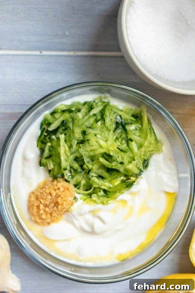 Recipe Tzatziki Sauce A bowl containing Greek yogurt, grated garlic, lemon juice, olive oil, and salt, with shredded cucumber ready to be added, for making Tzatziki sauce.