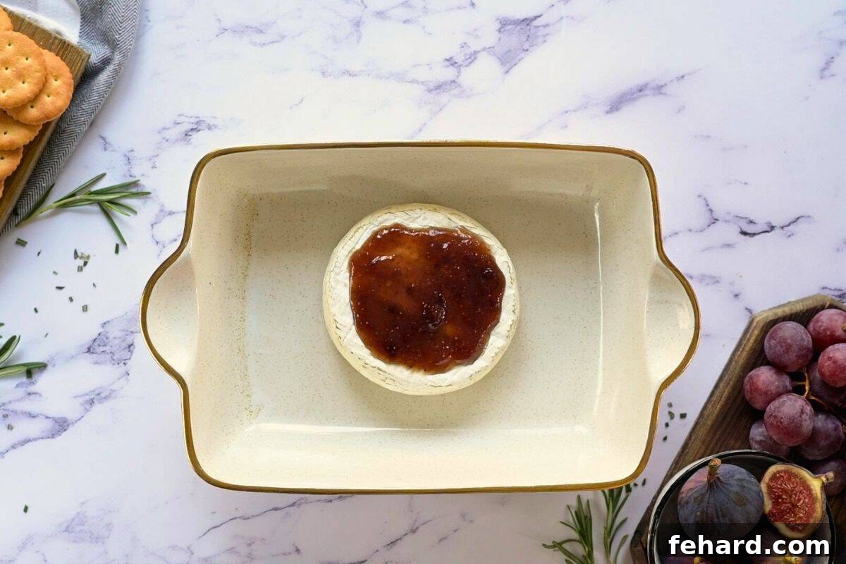 A baked wheel of brie cheese in a small rectangular baking dish, topped with fig jam and garnished with fresh figs and rosemary sprigs.