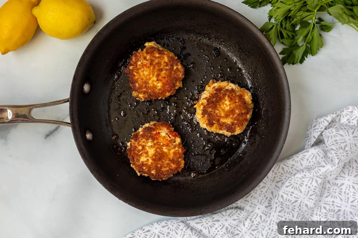 Three golden-brown crab cakes sizzling in a non-stick frying pan with hot oil, showcasing their perfectly crispy exteriors.