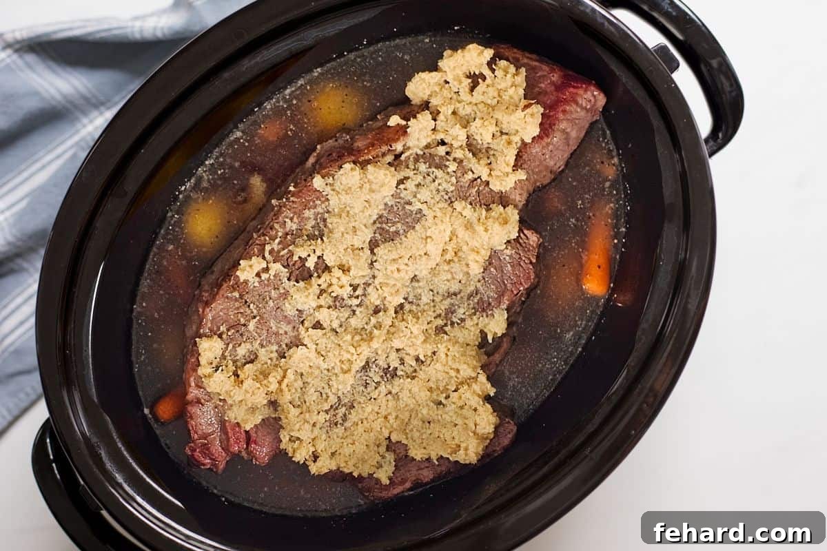A slow cooker filled with beef stock, layered with potatoes and carrots, and topped with a seared chuck roast smeared with prepared horseradish.