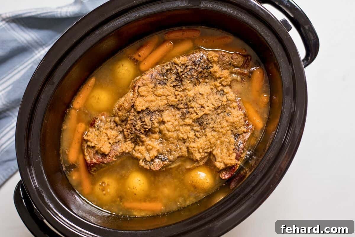 Slow-cooked chuck roast in a slow cooker, surrounded by rich beef broth, tender baby potatoes, and sweet baby carrots.
