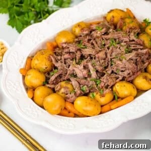 Shredded crockpot roast and potatoes and carrots on white platter next to parsley, horseradish, and gold serving spoons.