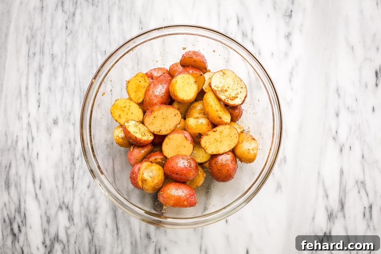 A close-up, overhead view of halved baby potatoes coated in a savory mixture of olive oil and spices, resting in a clear glass bowl, perfectly seasoned and ready for air frying.