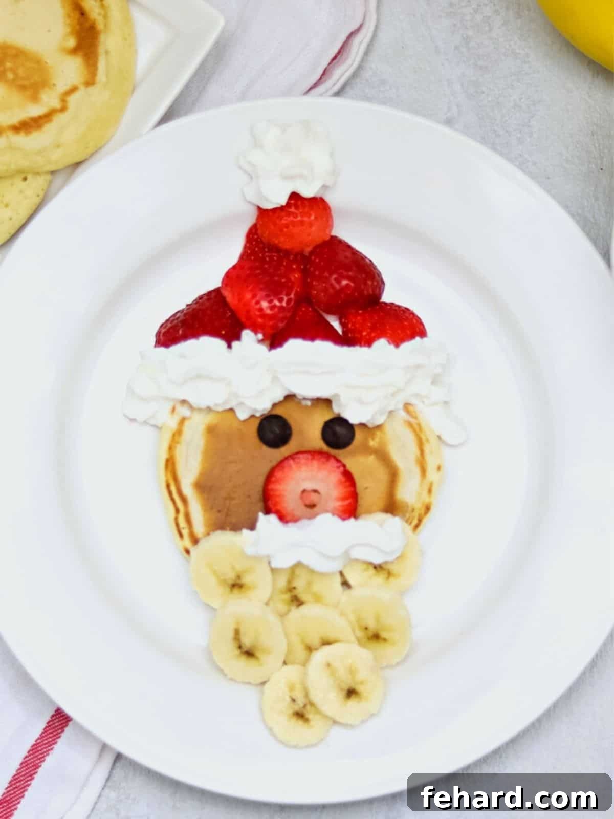 A beautifully arranged Santa shaped pancake on a white plate, featuring a fluffy pancake for the face, vibrant red strawberries for his hat, sliced bananas creating a fluffy white beard, and whipped cream details for the trim and mustache. This festive breakfast is garnished with chocolate chip eyes, ready for a magical Christmas morning.