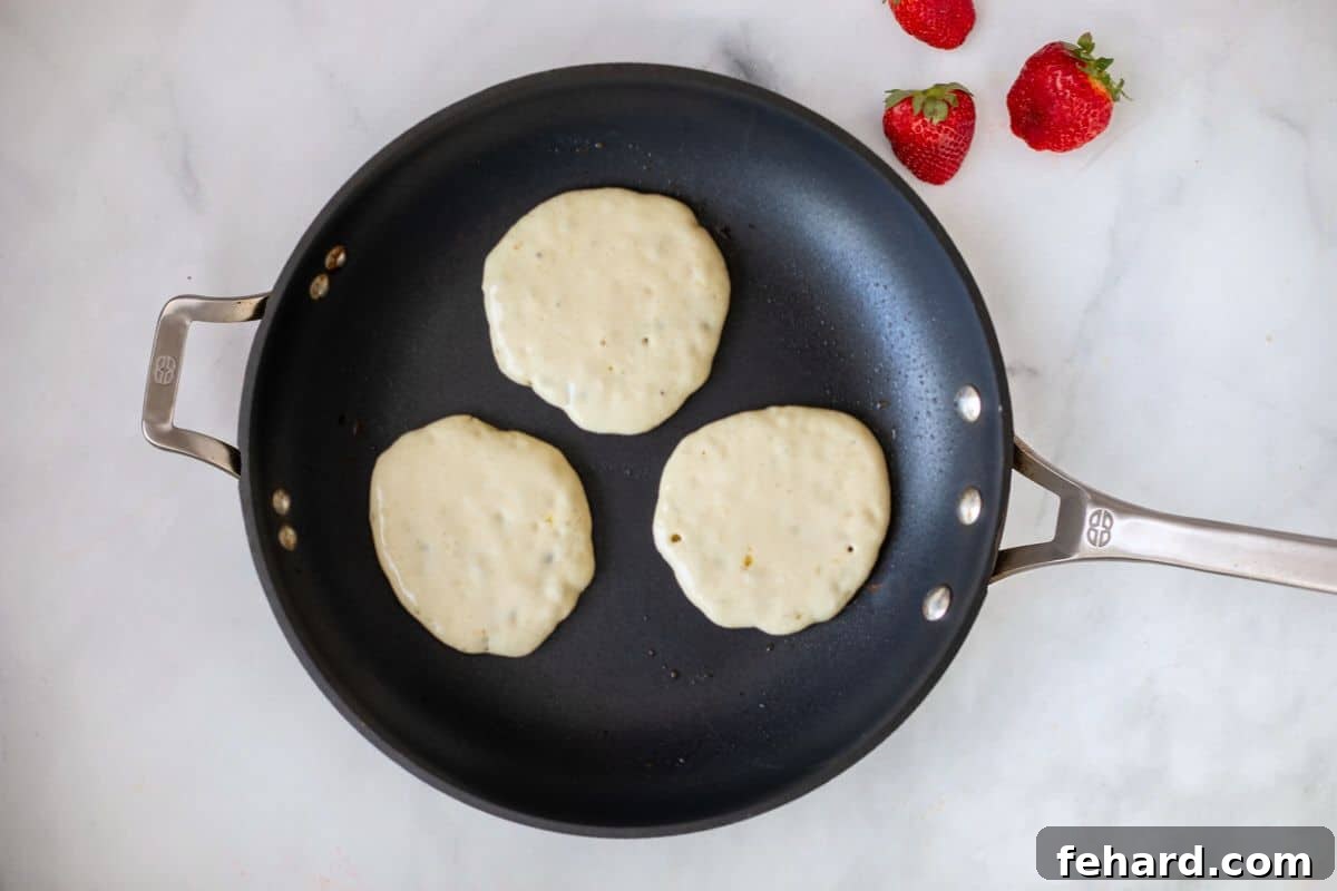 Three perfectly golden-brown pancakes sizzling gently in a nonstick skillet, serving as the essential base for building delightful Santa Pancakes. The scene captures the cooking process before decoration.