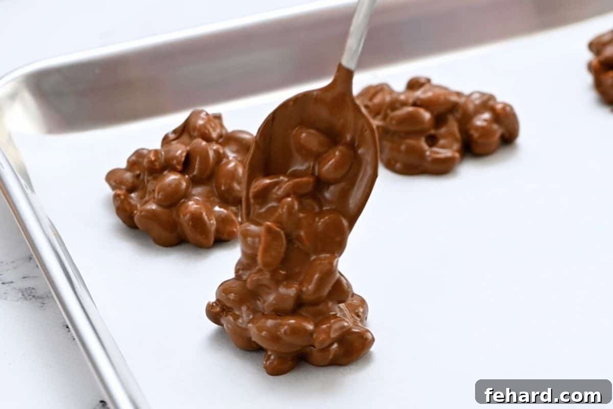 Scooping Easy Nut Clusters from Slow Cooker Peanuts generously coated in a rich chocolate mixture, being expertly spooned onto a parchment-lined baking tray directly from the crockpot.