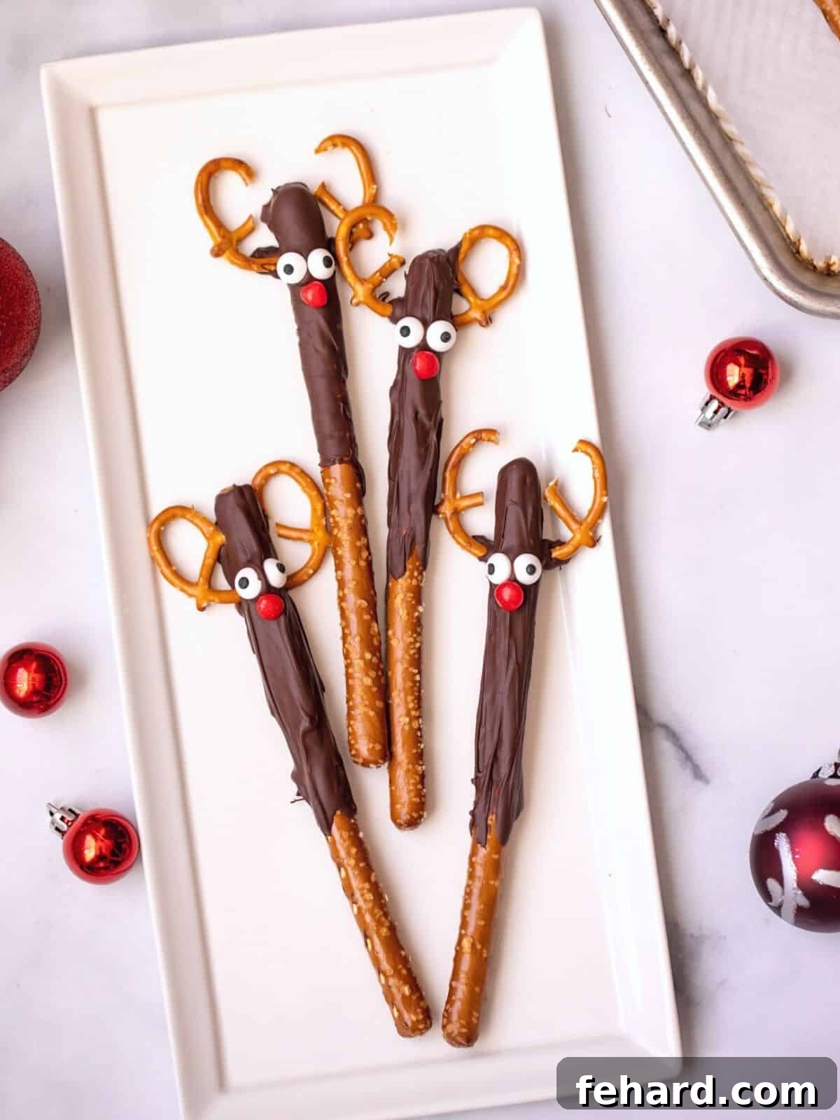 Four chocolate-covered pretzel rods decorated to resemble Rudolph the Red-Nosed Reindeer, artfully arranged on a pristine white platter, ready for a festive gathering.