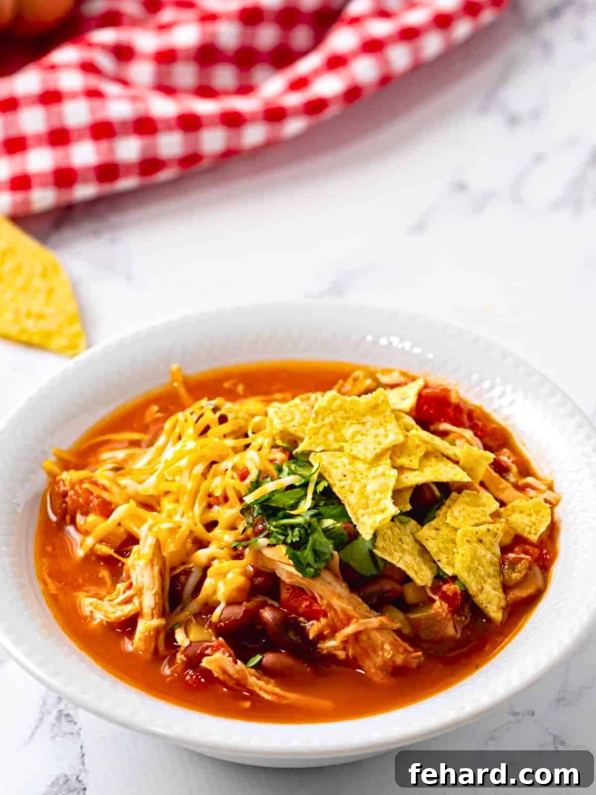 Slow cooker chicken chili with tomatoes and red beans served in white bowl topped with shredded cheese, minced cilantro, and tortilla chips.