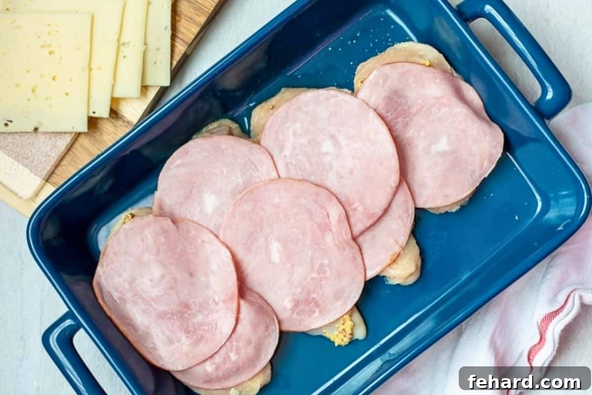 Chicken breasts brushed with Dijon topped with sliced ham in blue baking dish.