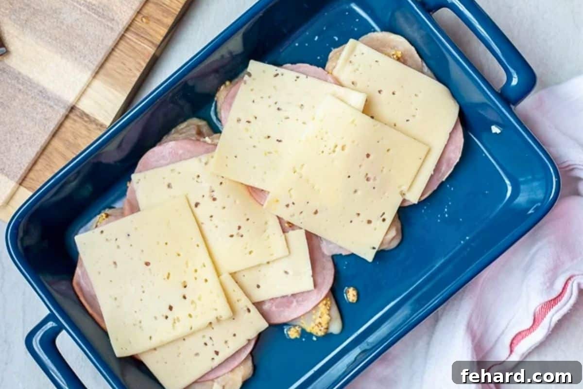 Chicken breast topped with ham and Swiss cheese in large blue baking dish.