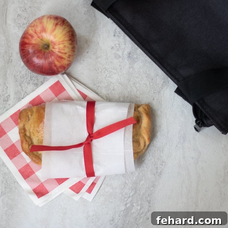 A ham and cheese hot pocket, neatly wrapped in parchment paper, next to a fresh apple, packed for lunch.