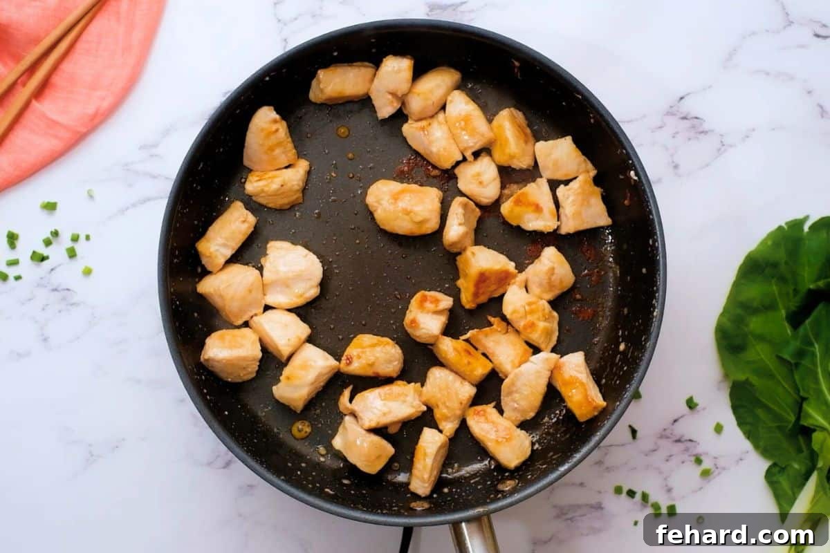 Close-up of browned chicken pieces sizzling in a hot sauté pan, preparing for a chicken and bok choy stir-fry.