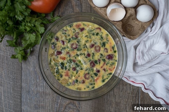 Mediterranean Spinach Frittata mixture, including eggs, spinach, tomatoes, olives, and feta, in a large mixing bowl, ready for baking.