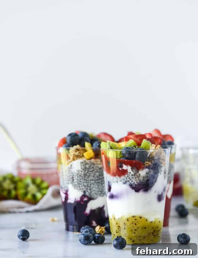Two vibrant Rainbow Syrup Bottomed Chia Pudding Parfaits showcased in clear glasses, beautifully topped with slices of kiwi, an assortment of fresh berries, and crunchy granola.
