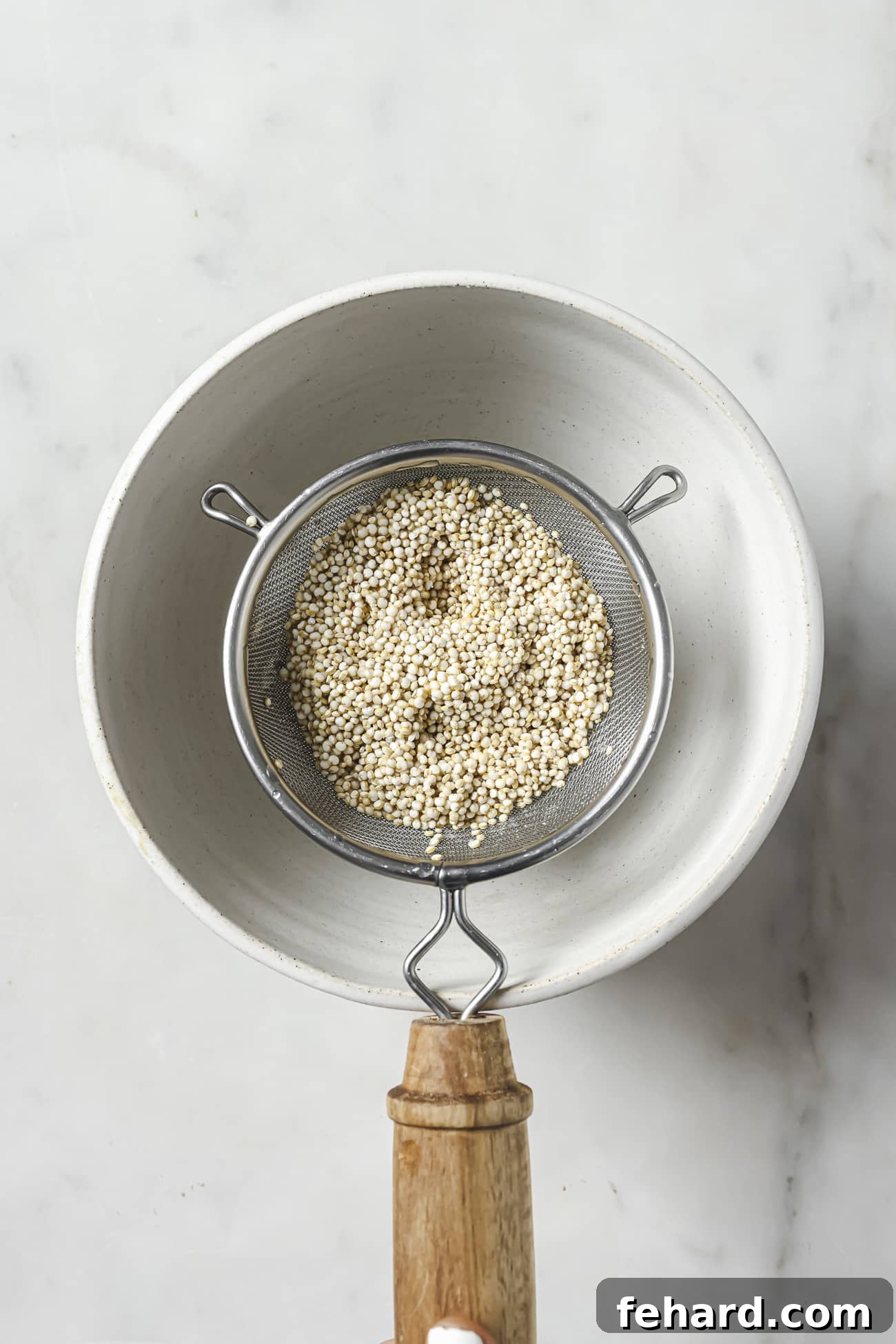 Uncooked quinoa in a fine mesh strainer being rinsed under cool running water, held over a white bowl to catch the excess liquid.
