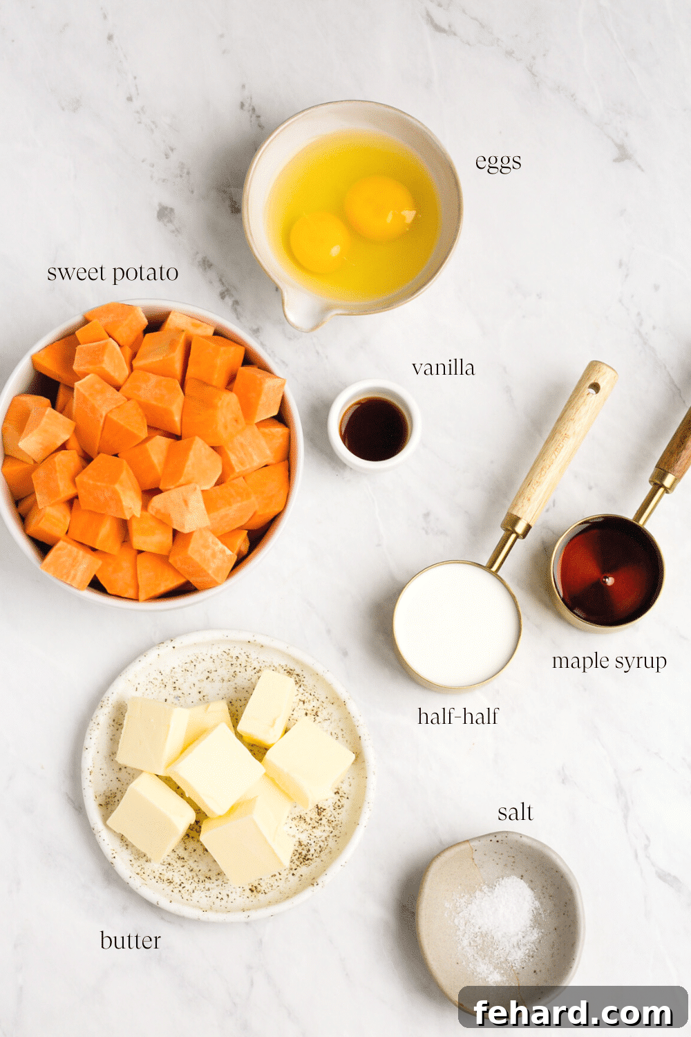 Overhead view of all the fresh ingredients for the sweet potato casserole, including whole sweet potatoes, butter, half-and-half, maple syrup, eggs, vanilla, and salt arranged in individual bowls.