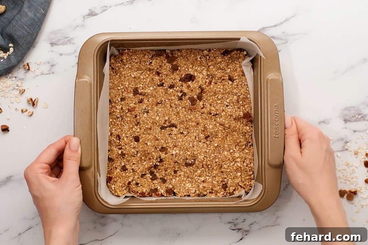 No Bake Granola Bars Pressed into Pan Smooth, compacted granola bar mixture pressed evenly into a square baking dish that has been carefully lined with parchment paper, indicating it's ready for the chilling process.