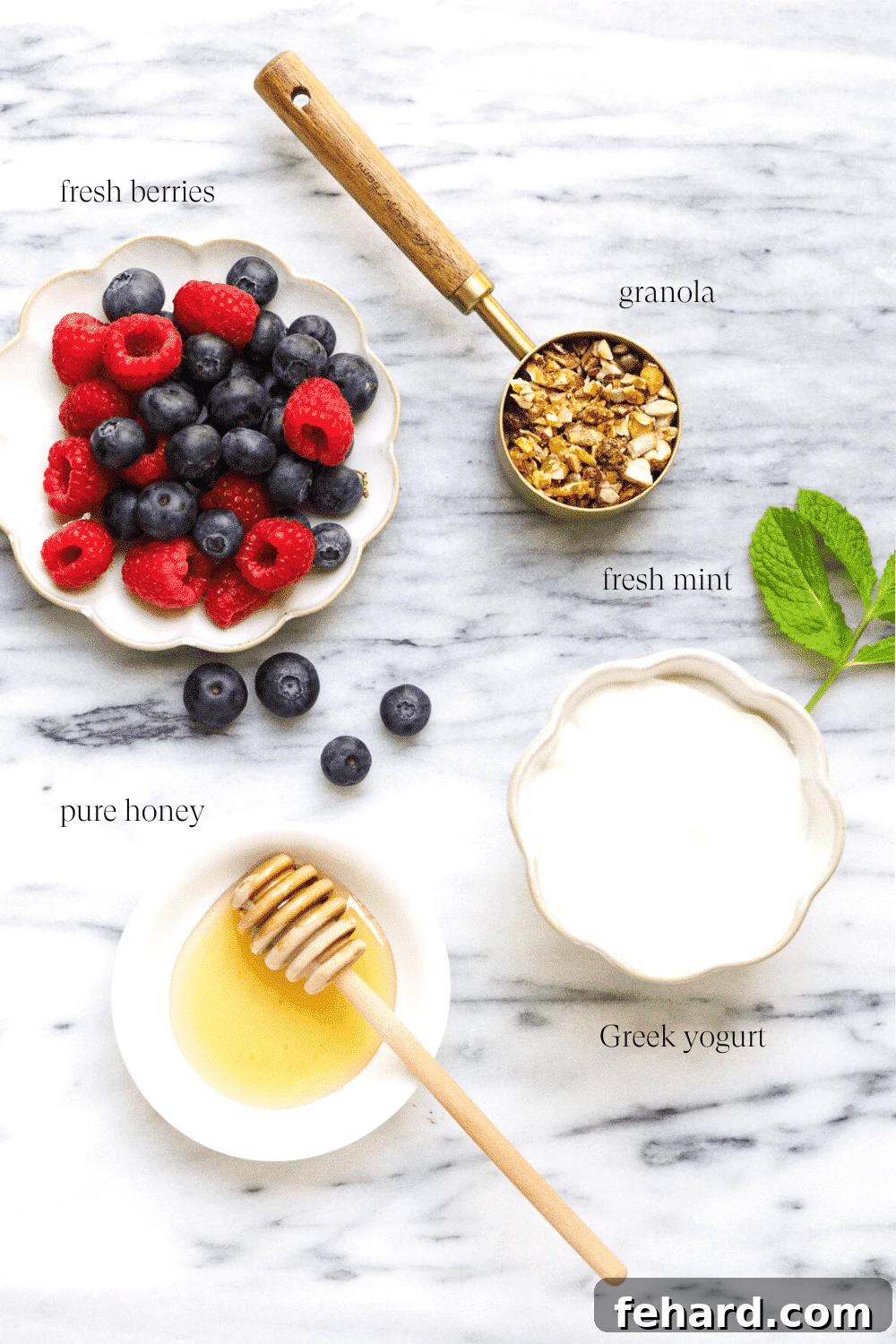 Overhead view of fresh ingredients laid out on a table, including Greek yogurt, various berries, granola, honey, and mint leaves, ready for making a yogurt parfait.