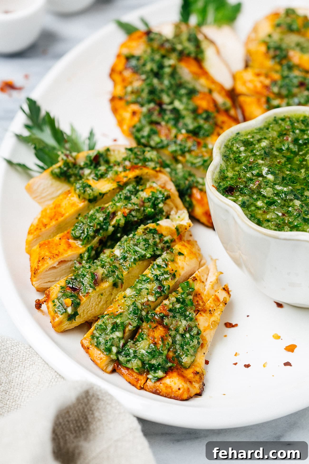 Close-up overhead shot of a single grilled chicken breast, beautifully charred, topped with vibrant green chimichurri sauce on a white plate.