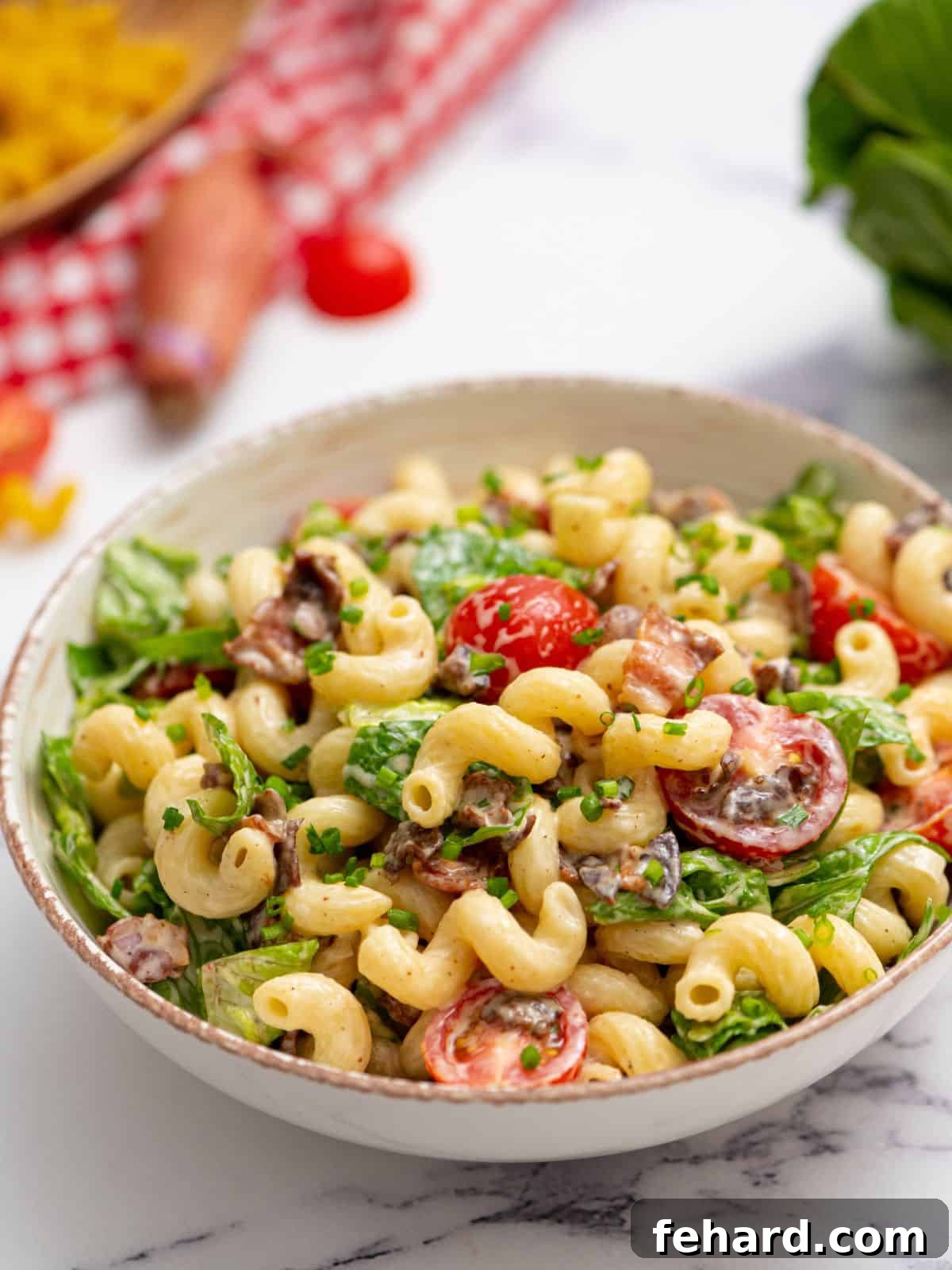 Creamy BLT Pasta Salad - Ultimate Recipe A bowl of vibrant, creamy BLT Pasta Salad featuring visible pieces of crispy bacon, bright red grape tomatoes, and fresh green lettuce, with additional grape tomatoes blurred in the background.