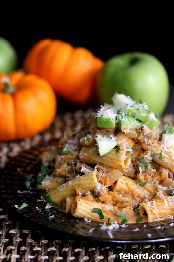 A black plate generously piled with rigatoni pasta bathed in pumpkin sauce, mixed with savory sausage pieces and crisp green apple slices.