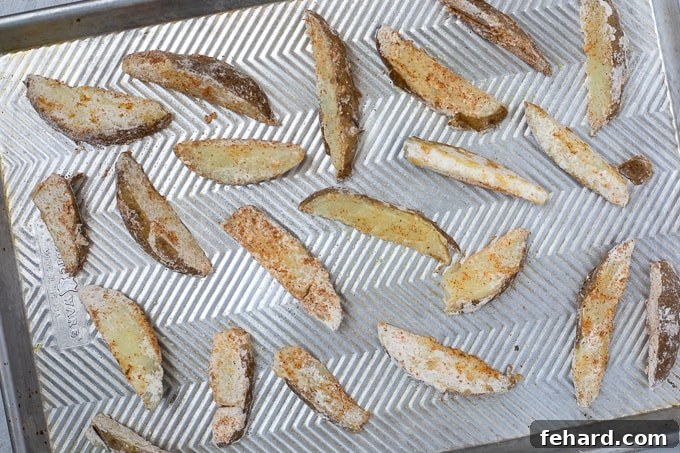 Seasoned potato wedges spread out in a single layer on a greased baking sheet, ready for the oven.