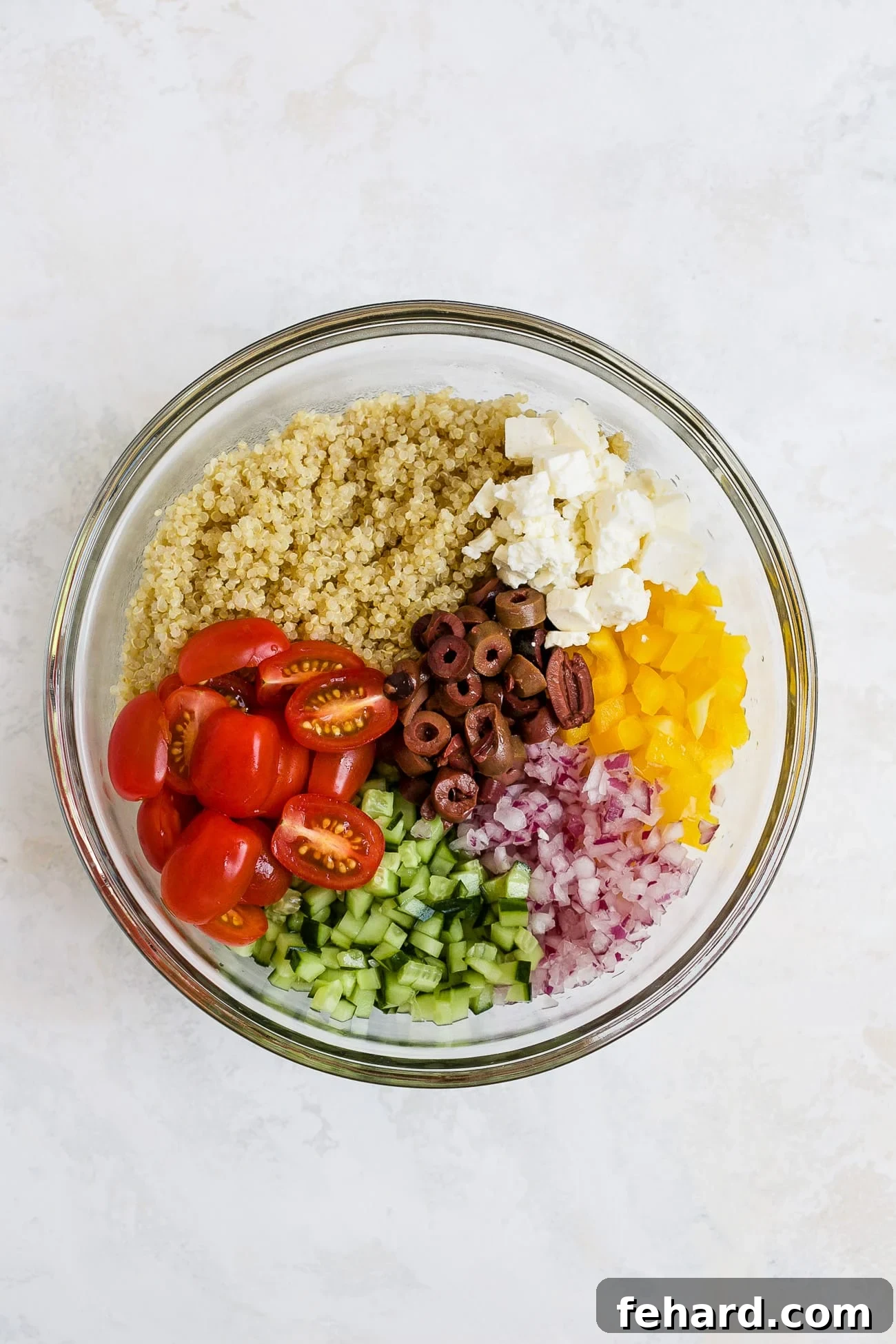 A large bowl filled with cooked quinoa, colorful cherry tomatoes, black olives, crisp cucumber, red onion, bell pepper and feta, all mixed together before dressing.
