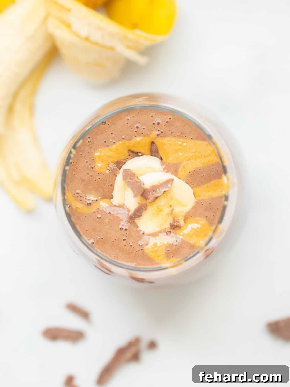 A stunning top-down view of a rich chocolate peanut butter smoothie served in a clear glass. The smoothie is elegantly topped with a swirl of creamy peanut butter, delicious chopped chocolate pieces, and perfectly sliced banana rounds, showcasing its appetizing presentation.