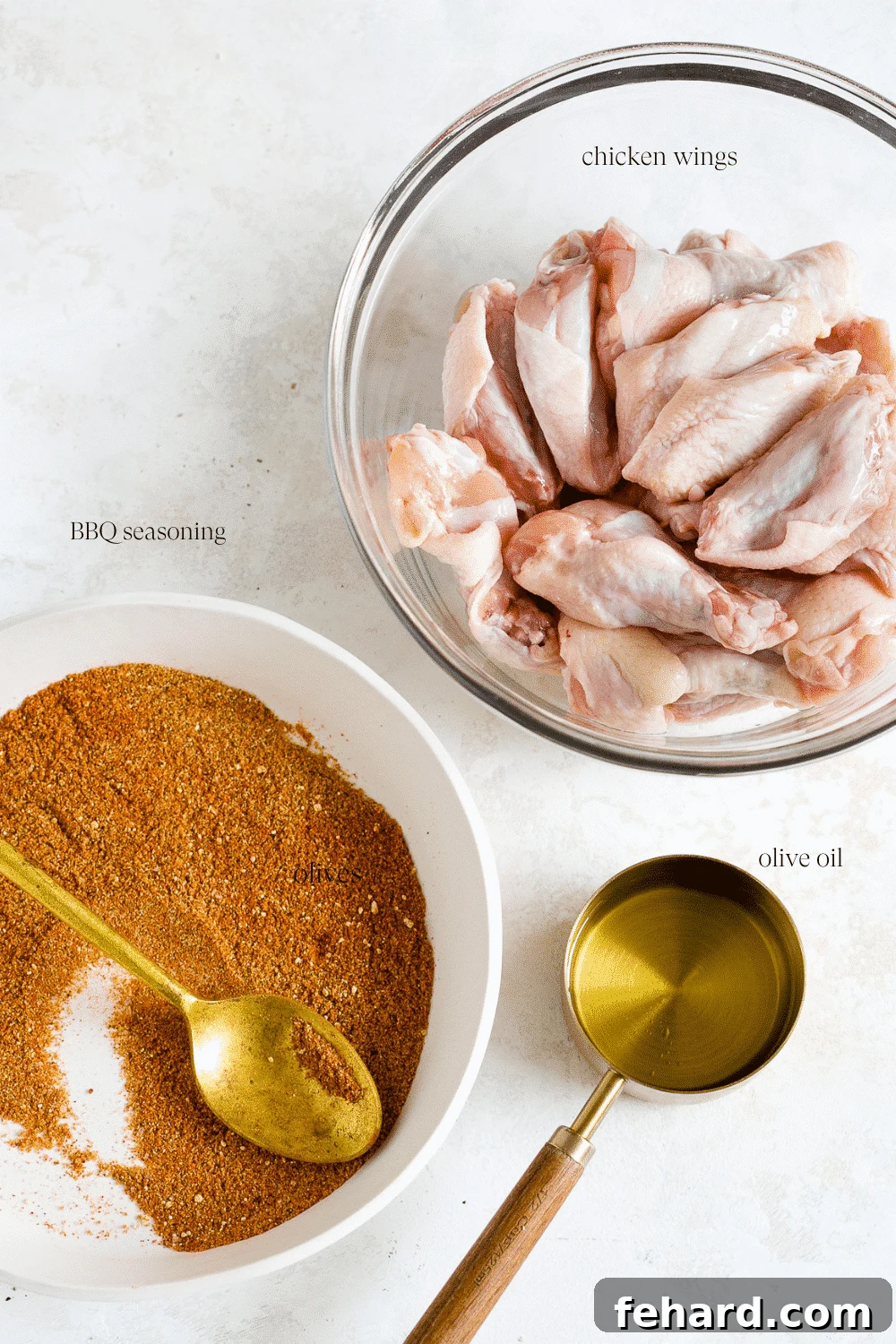 Raw chicken wings in a clear glass bowl, accompanied by a white plate of barbecue seasoning and a measuring cup