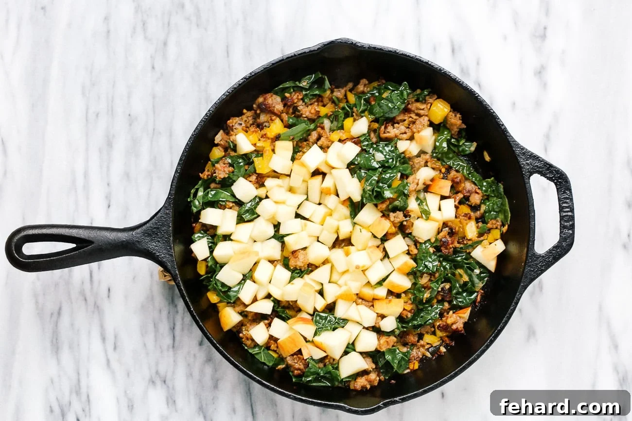 Chopped kale, diced apple, and fresh sage added to the skillet containing browned sausage and vegetables, mid-stir.