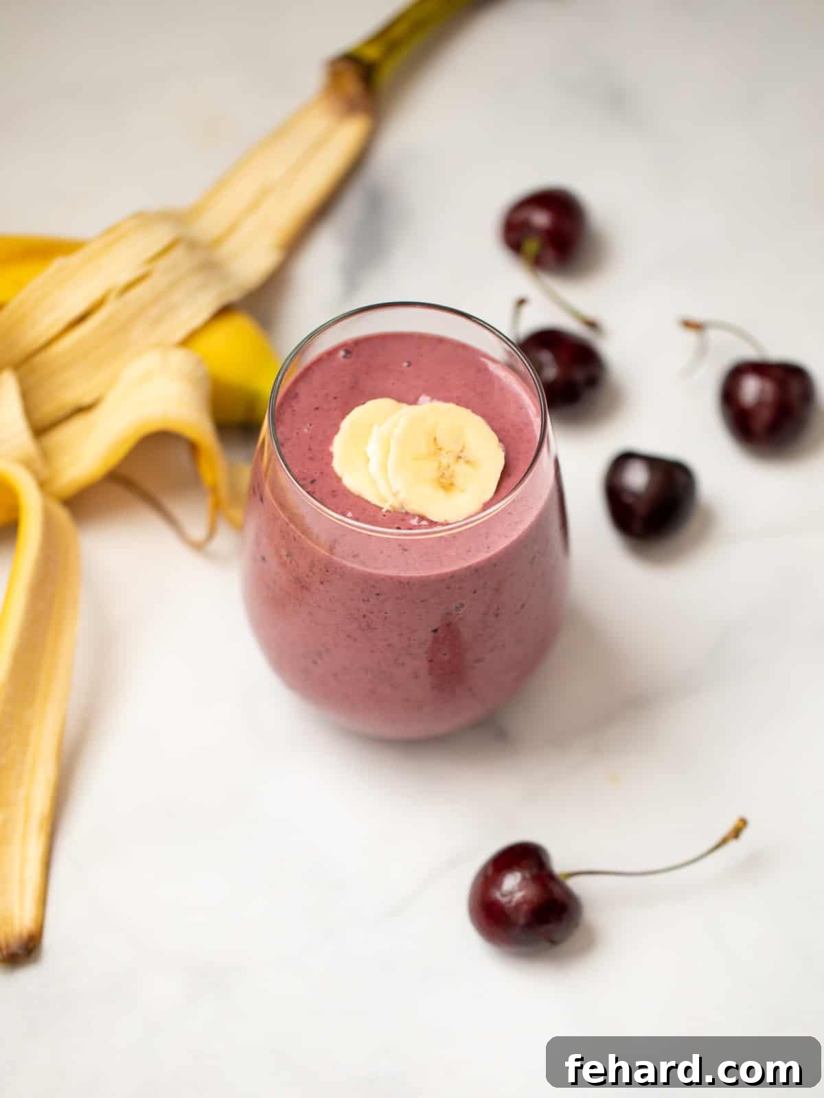 Delicious Cherry Smoothie Recipe Vibrantly red Cherry Smoothie in a clear glass, garnished with a fresh cherry, alongside a scattering of fresh cherries and a ripe banana, set against a rustic background. This image perfectly captures the fresh and inviting nature of the smoothie, ideal for a healthy and quick meal.