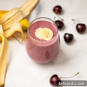 Close-up of a Cherry Smoothie in a clear glass, beautifully garnished with banana slices and a few fresh cherries, with more fresh cherries visible in the background.