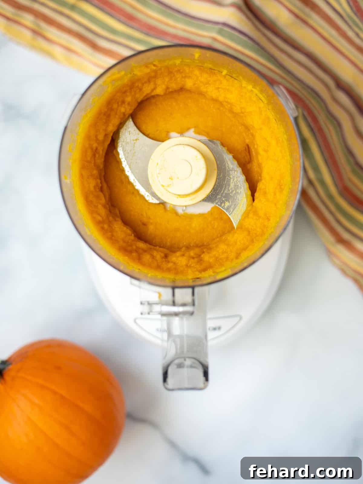 Homemade Pumpkin Puree: Easy Oven and Instant Pot Recipe Food processor with homemade pumpkin puree inside bowl, with a raw sugar pumpkin and a cinnamon stick on the side, highlighting the freshness and ease of the process.
