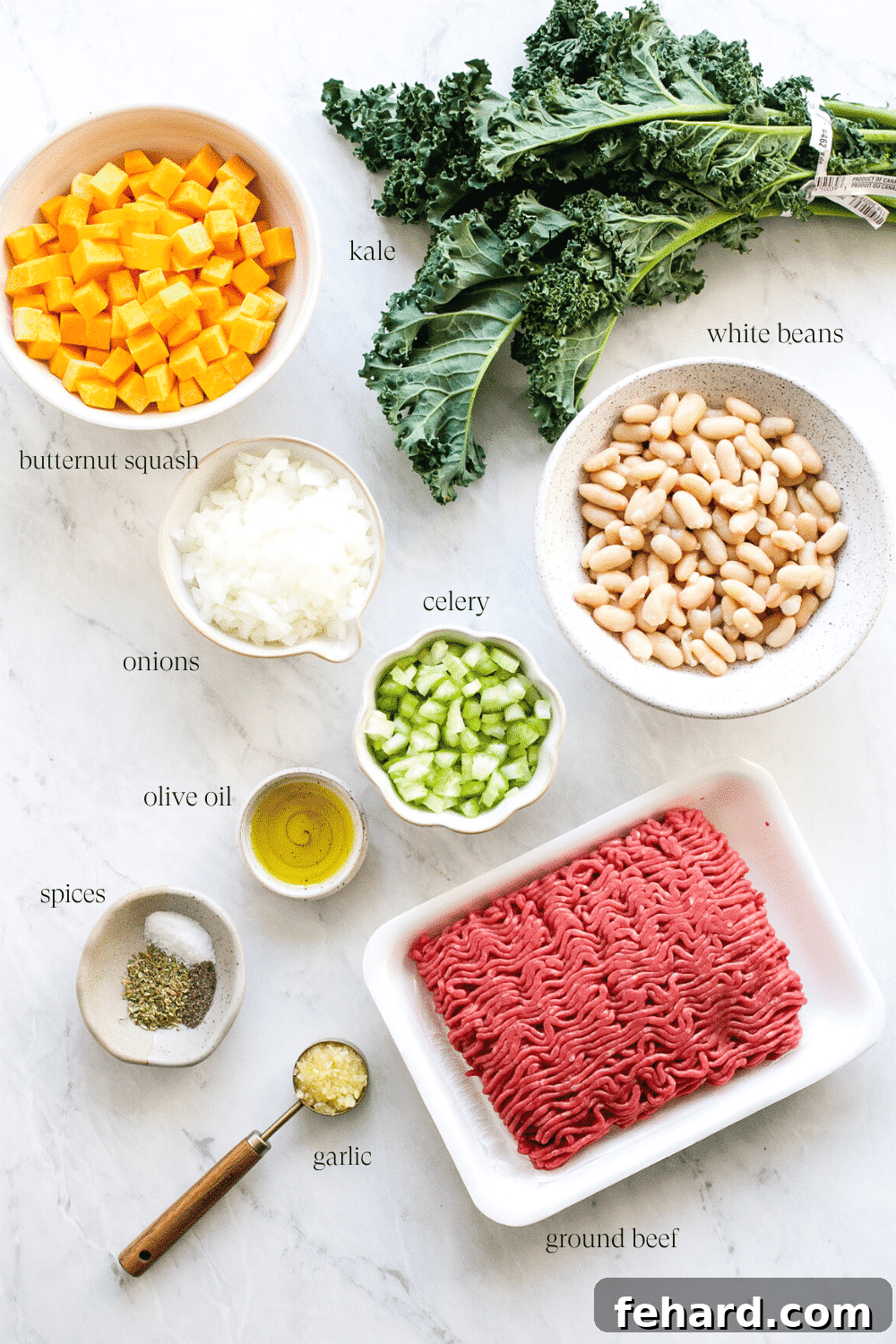 Wholesome White Bean and Kale Pot 3 A collection of fresh ingredients for white bean and kale soup, including butternut squash, kale, onions, celery, garlic, ground beef, and white beans, arranged on a kitchen countertop.
