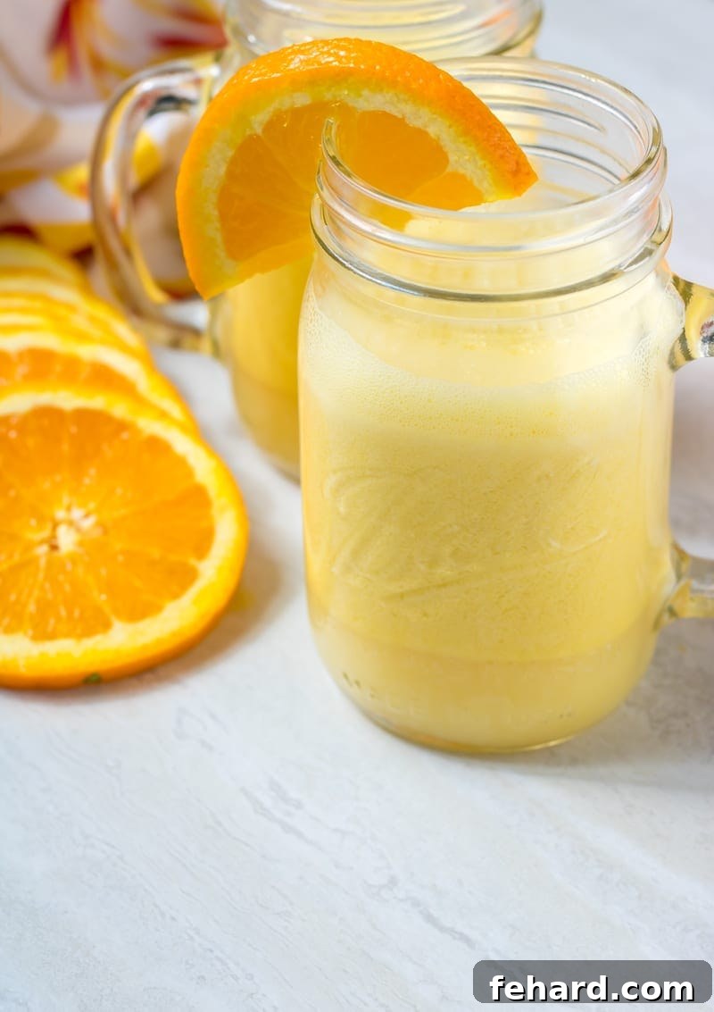 A tall, elegant glass cup filled with a frothy homemade Orange Julius, garnished with a vibrant fresh orange slice, perfectly positioned on a clean, light-colored surface.