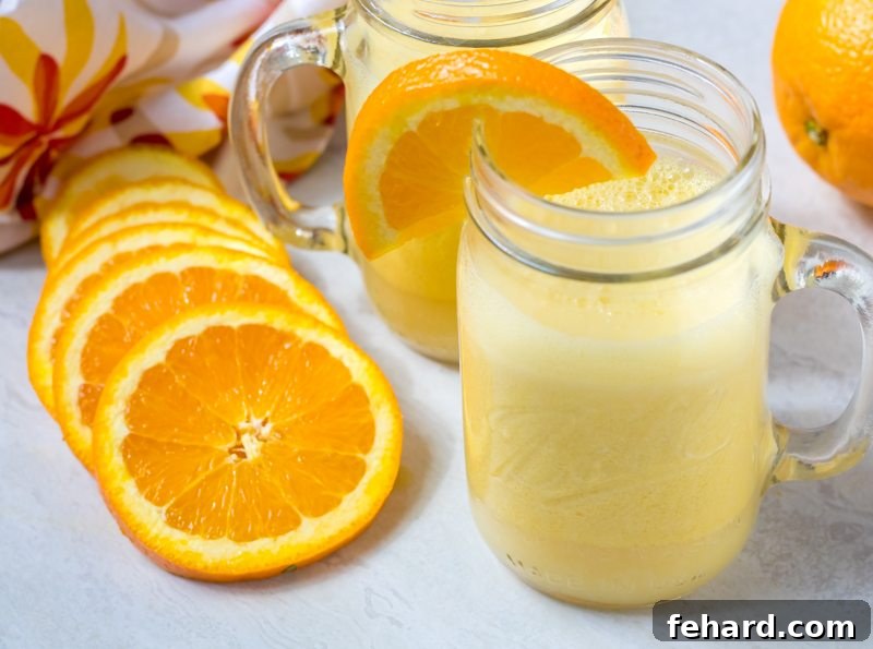A beautifully blended homemade Orange Julius in a clear glass, set on a rustic wooden counter. Fresh orange slices are scattered around, and a modern blender is subtly visible in the soft-focus background, highlighting the ease of preparation.