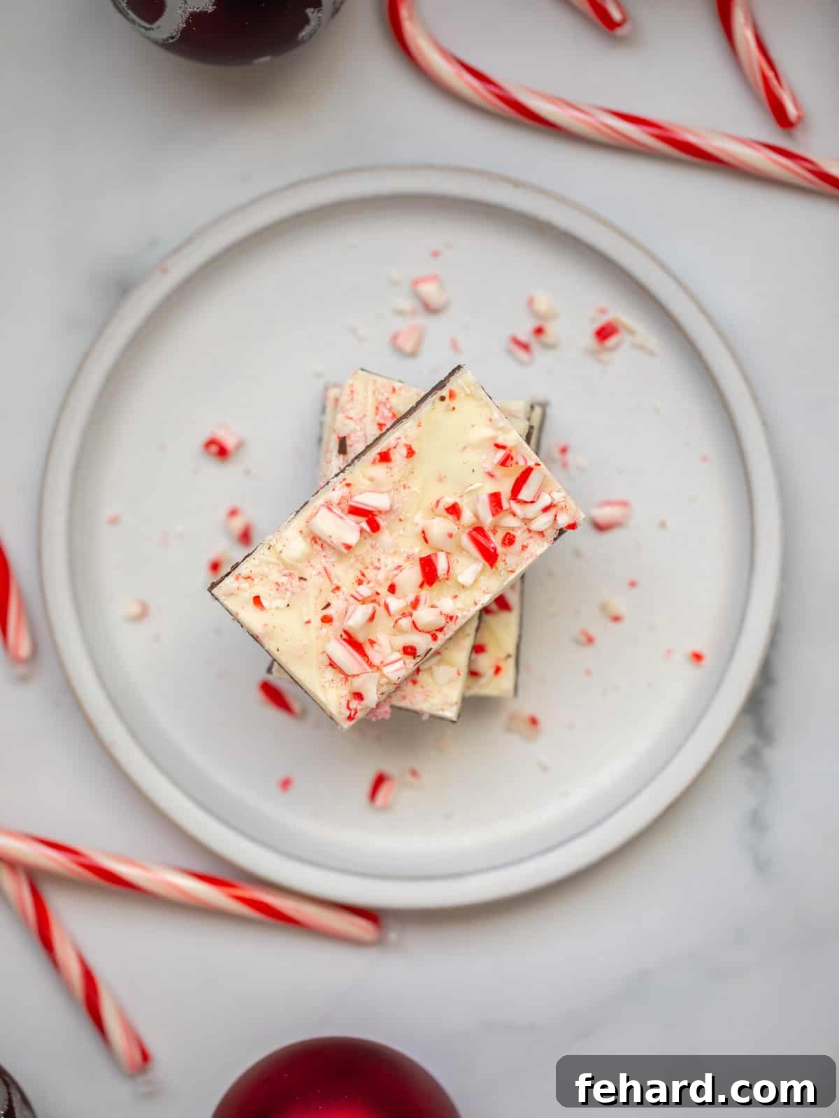 Homemade Peppermint Bark cut into rectangles on a plate next to Christmas ornaments and candy canes, ready to be enjoyed.